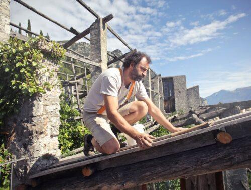 Side view of male builder in summer wear reconstructing rooftop and sitting on wooden beams of aged building on background of scenic landscape in sunny day