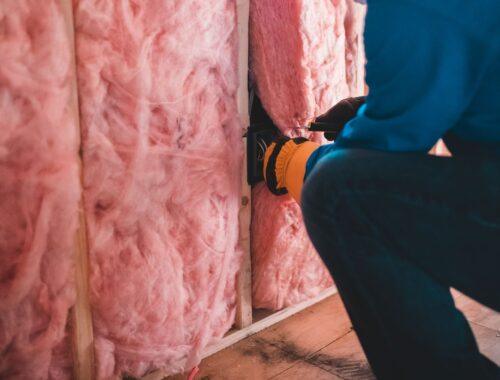 Crop unrecognizable worker in gloves sitting on haunches and insulating with pink stone wool